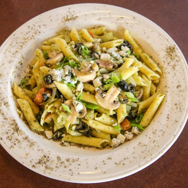 Penne pasta with pesto, mushrooms, black olives, tomatoes, and crumbled cheese on a white plate.